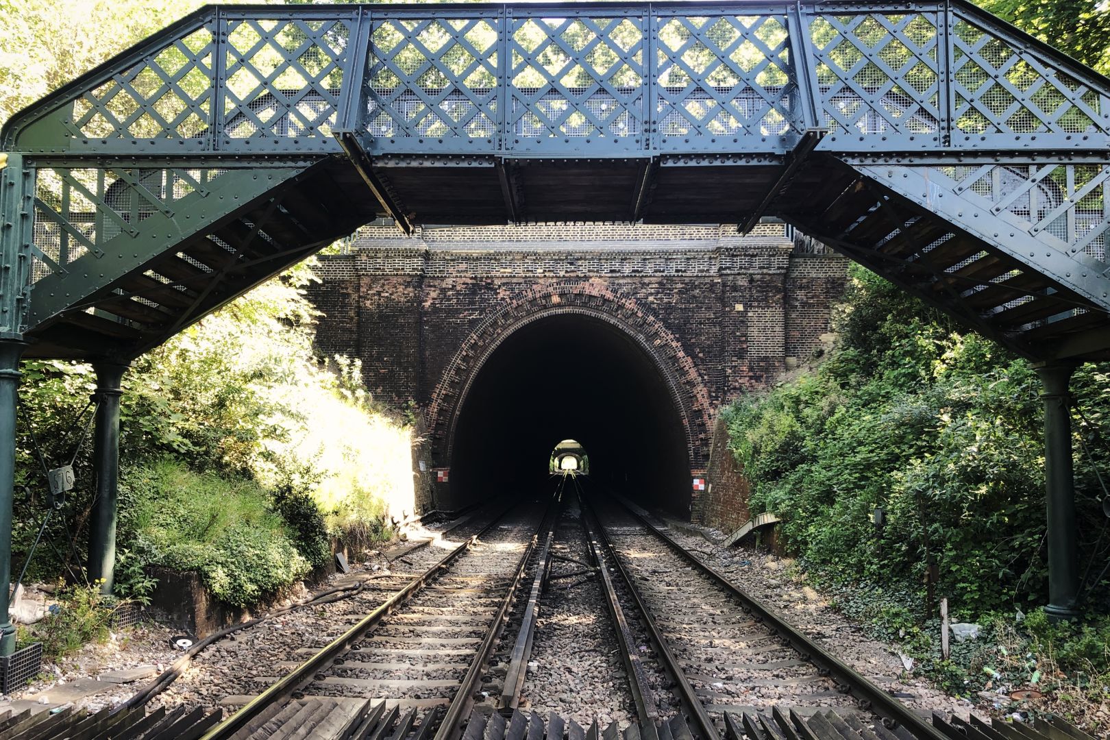 Two railway tracks disappear into a tunnel; in the far distance some light is visible where the line emerges and then perhaps enters another tunnel soon afterwards. On this side of the tunnel the tracks are surrounded by bushes, and a Victorian ironwork bridge, painted dark green, stretches over the line to allow pedestrians to cross when the crossing is closed to road traffic. In the extreme foreground, the very edge of the level crossing is just visible.