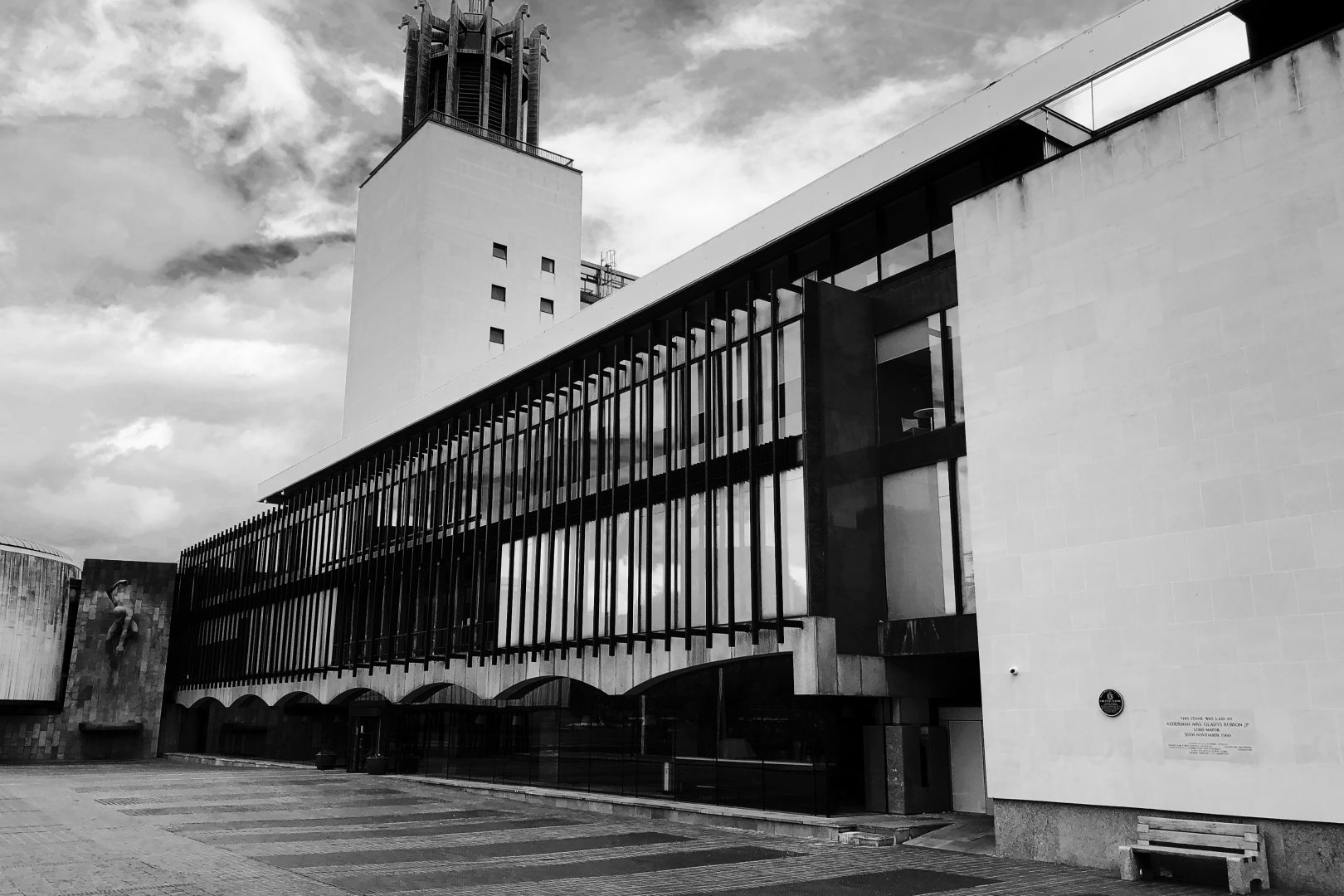 A long modernist building with glass and concrete sides. The ground floor has long concrete arches; the upper floors have windows with decorative metalwork. At the far end is a tower: a sort of modernist interpretation of a bell tower. To the left of the tower, the building starts to extend out in an L-shape, and on this part of the building is a fountain with a statue of a bearded mythological figure, larger than human height.