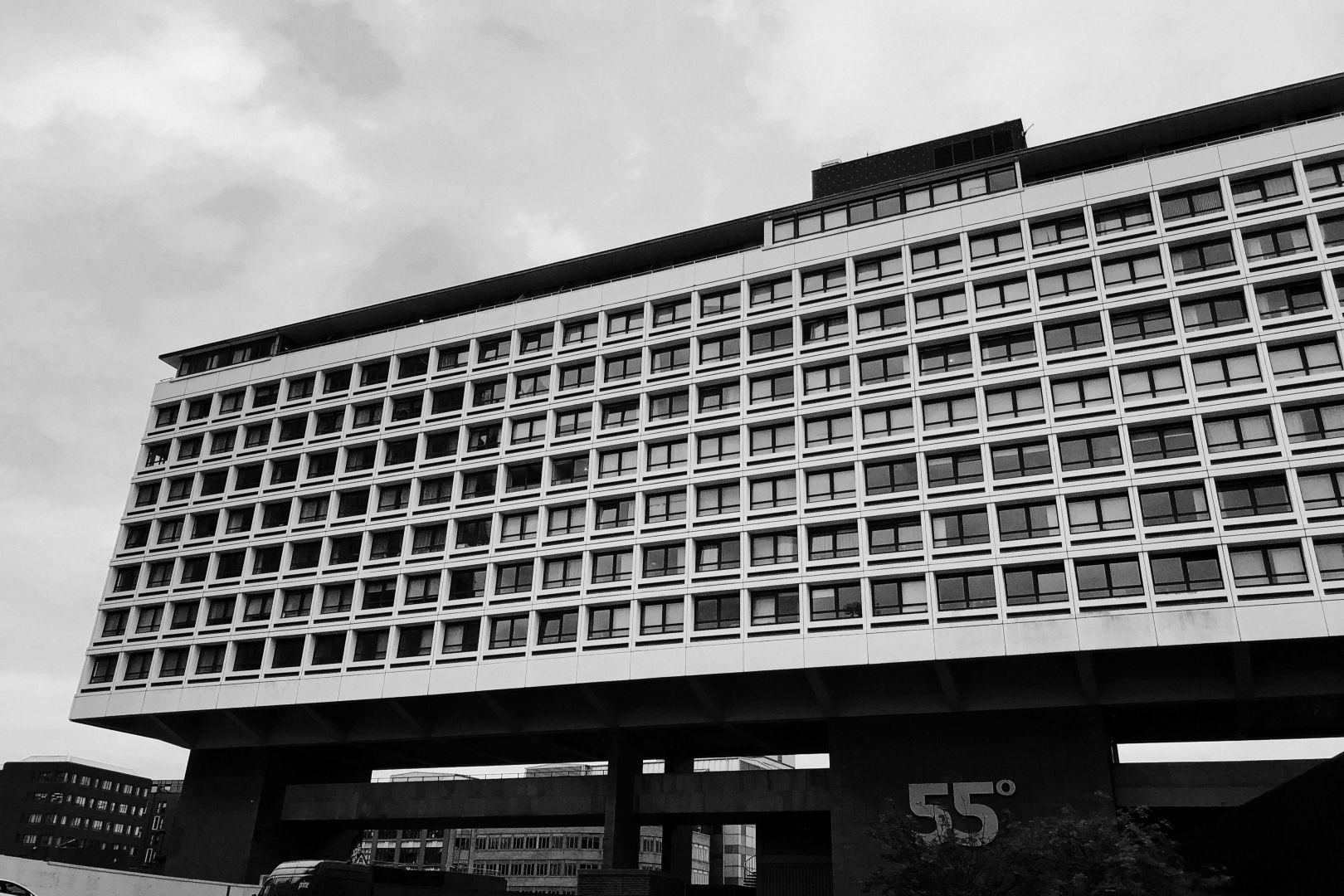 A long white modern building with many windows. It’s supported on dark concrete pylons, several storeys high, with a large sign reading “55°”.