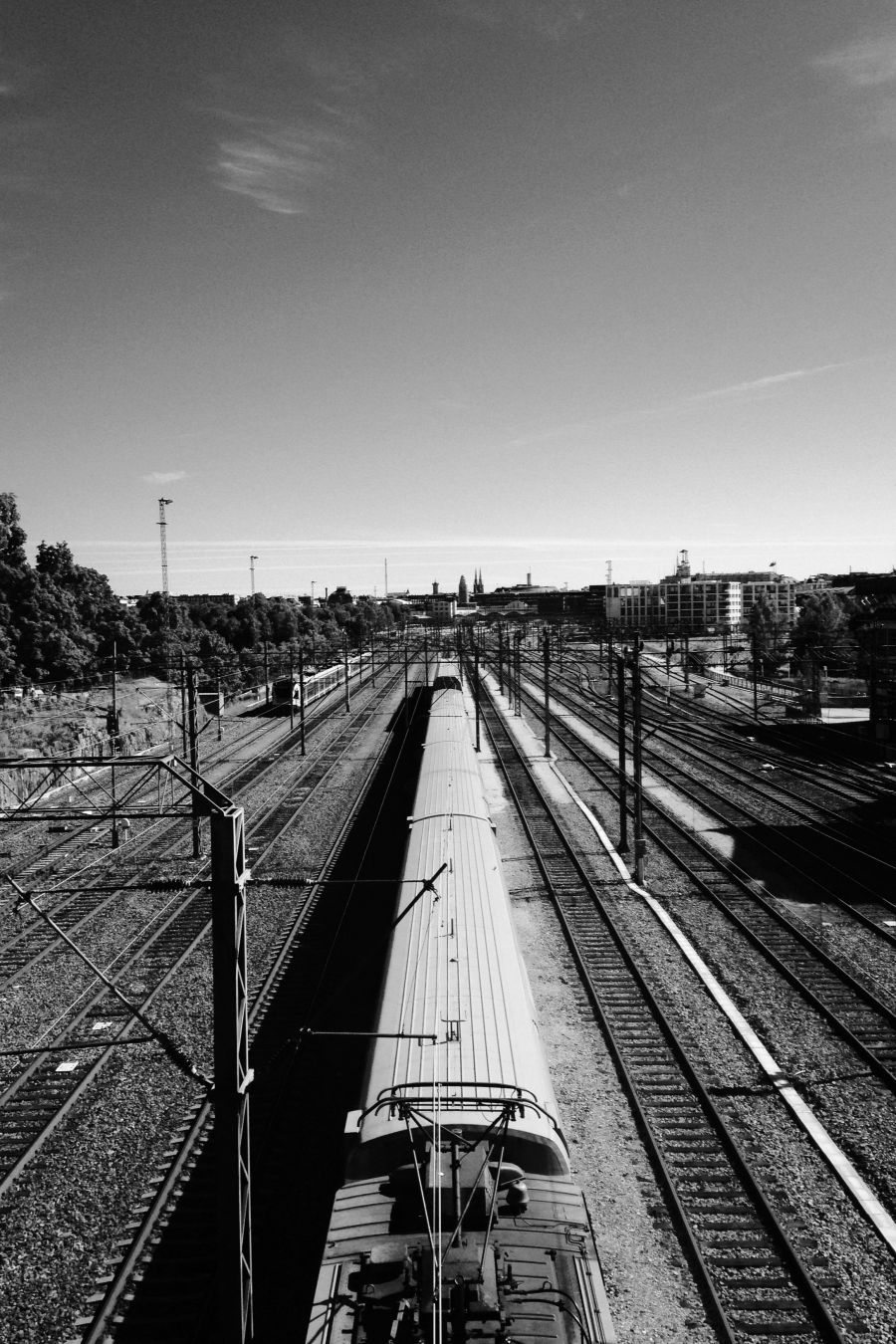 Many parallel railway lines viewed from above (on a bridge), converging in the distance; at least ten or twelve, though on the right several of them split and merge on way that makes them hard to count. On the central line is a train (modern, electric), extending under the viewer’s feet; on the leftmost line, further away in the distance, is another, smaller train. All around are the paraphernalia of railways: pylons, cables. On the leftmost side the lines are edged by a cliff that rises above them and is topped by trees. In the distance some buildings are visible: the tower of the central railway station and the spires of a church, among other things, poke out above the mid-rise office blocks and shops of the city.