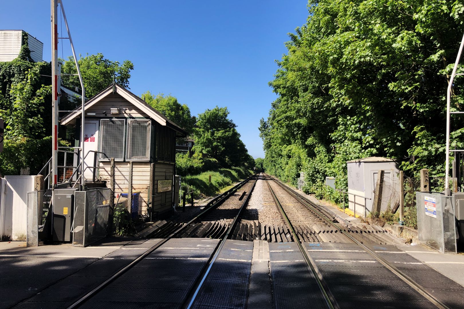 Two railway tracks stretch away into the distance, perfectly straight, and lined with trees on either side. In the foreground a road crosses the line at a level crossing; on both sides the gate is open to allow traffic through. On the left is a small hut, somewhat run-down. It has metal grills over the window and modern safety signs stuck to the door, but just about readable on the side is the name of the crossing — “Charlton Lane” — in an old-fashioned font (1960s, perhaps).