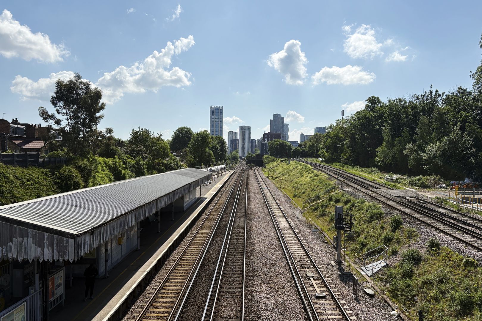 Railway lines stretching into the distance. A run-down station building on the left, Victorian architecture with flaking paint on the wooden canopy. On the right, an embankment carrying the express lines past the station. In the distance are some tall buildings, and, barely visible at their base, the next station on the line.