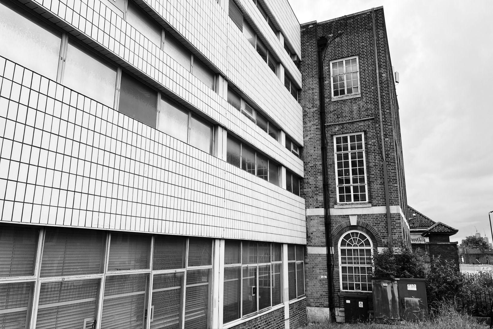 A black-and-white photograph of a slightly run-down building, taken at an angle of the façade. The left two-thirds are mid/late twentieth-century: white tiles, plate windows running the length of the building, blinds visible through them. At the right-hand end the building looks older, maybe early twentieth century: brick, with large vertical windows made up of smaller panes.

In front of the building are some electric cabinets partly overgrown with long grass and some sort of shrub. Just visible next to it, further along the street, is the roof of another abandoned building, with a security camera and spikes on the wall.

In the distance on the far right hand side, over the tree line some towers and cranes can be seen.